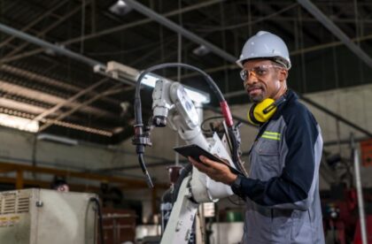 A man wearing a hard hat, safety glasses, and earmuffs holding a tablet beside a robotic arm in a factory.