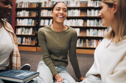 Three women sitting on a library floor, talking and smiling near bookshelves with a book in the foreground.