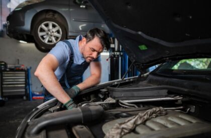 A man wearing overalls and gloves working under the open hood of a car inside an auto repair shop.