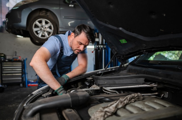 A man wearing overalls and gloves working under the open hood of a car inside an auto repair shop.