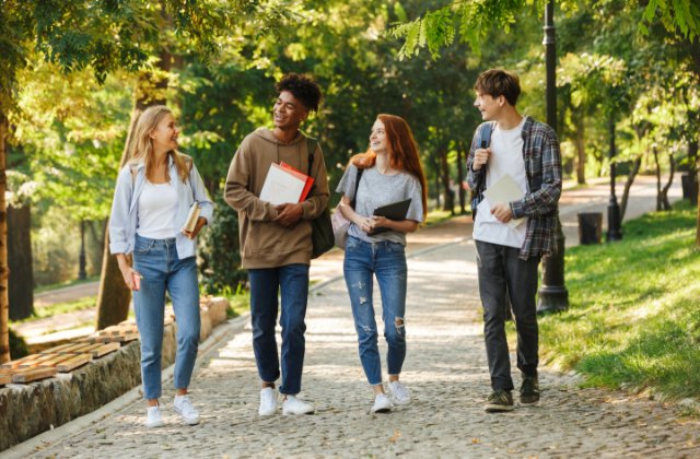 Four young adults carrying books and folders while walking together on a tree-lined path in daylight.