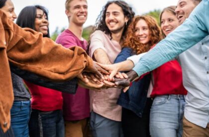 A group of smiling young adults standing outdoors. They put their hands together in the center of their circle.