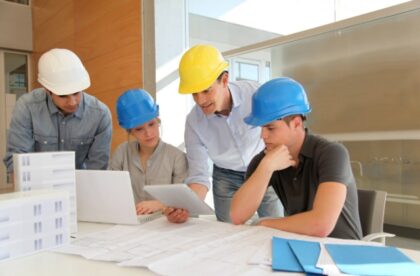 A close-up of four people wearing hard hats staring at a tablet beside architectural plans and a building model.