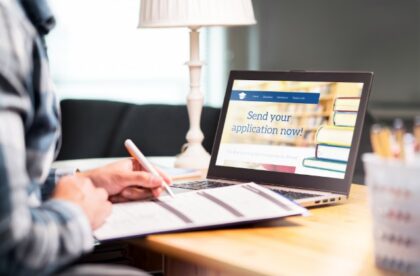 Person writing on paperwork beside a laptop displaying a college application website, with books and a lamp nearby.