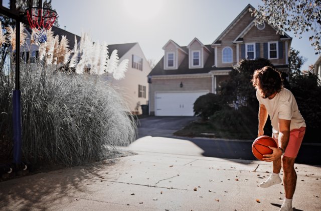 A close-up of a man wearing orange shorts, dribbling a basketball on a suburban driveway in front of a house.