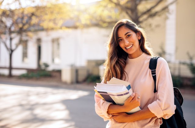 Young woman in a pale pink sweatshirt carries books and a black backpack while smiling on a tree-lined campus.