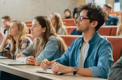 College-aged adults sit in an auditorium leaning on a table that stretches in front of the chairs. They look forward.
