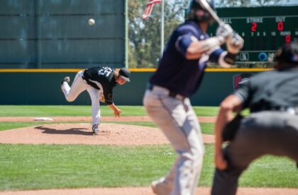 A view from behind home plate as a pitcher in a black jersey pitches to a batter in a blue jersey on a sunny day.