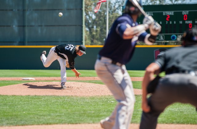 A view from behind home plate as a pitcher in a black jersey pitches to a batter in a blue jersey on a sunny day.