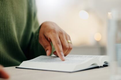 A person wearing a green sweater points to a line of text in a book. The words are arranged in two columns on the page.