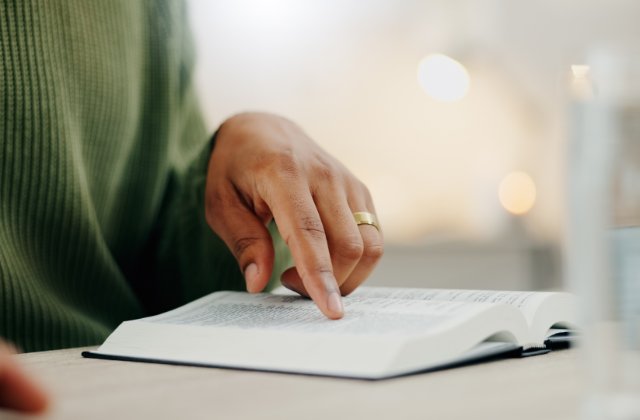 A person wearing a green sweater points to a line of text in a book. The words are arranged in two columns on the page.